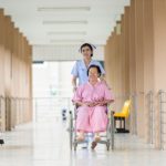 Nurse pushing an older woman in a wheelchair along an outdoor tiled corridor